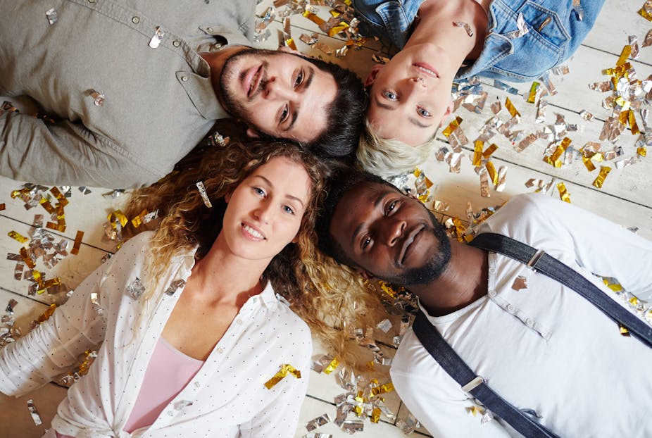 Empat anak muda yang berbaring dengan kepala menyatu di lantai, dengan confetti perak dan emas yang ditaburkan di atas mereka
