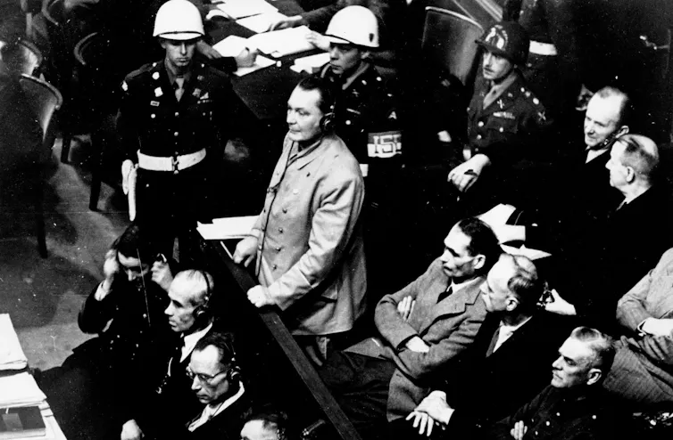 A black-and-white photo shows a dark-haired man standing in the prisoner's dock surrounded by armed guards and men in suits sitting near him.