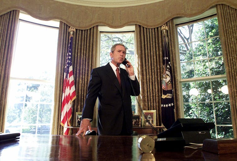 A man in a suit stands in an ornate office behind a large desk talking on the phone. Gold curtains and the American flag are behind him.