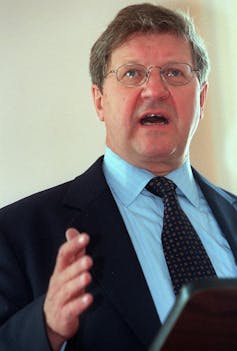 A man with glasses wearing a dark suit speaks from behind a lectern.
