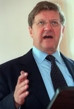 A man with glasses wearing a dark suit speaks from behind a lectern.