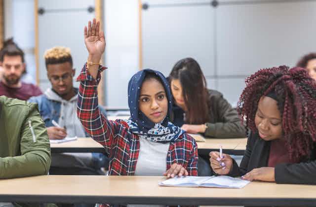 A student wearing a hijab has her hand raised for a question in a classroom.