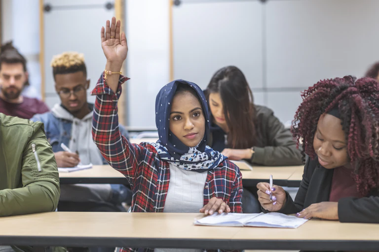 A student wearing a hijab has her hand raised for a question in a classroom.
