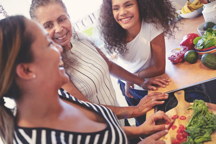 Three generations of First Nations people are standing at a kitchen bench together preparing vegetables and they're all smiling.