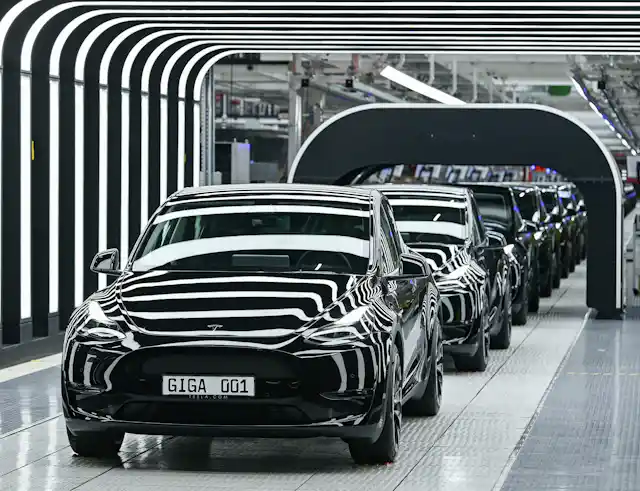 Several Tesla Model Y electric vehicles stand on a conveyor belt at the opening of a Tesla Gigafactory. Lighting creates a striped effect as the vehicles pass under a black arch.