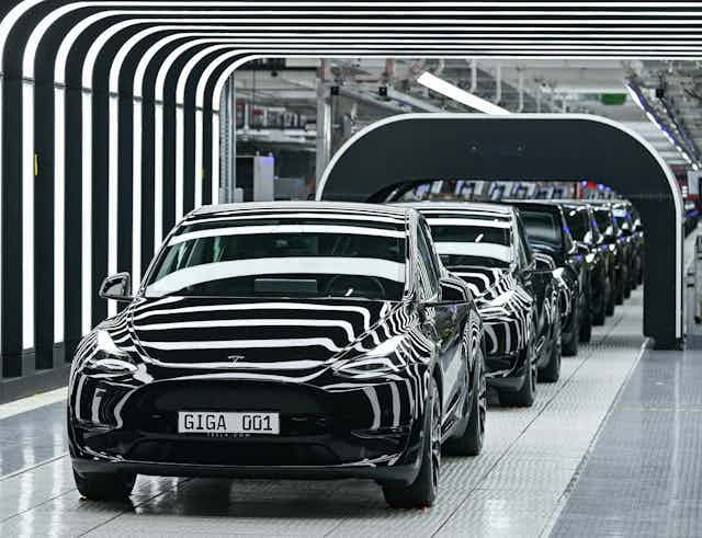  Several Tesla Model Y electric vehicles stand on a conveyor belt at the opening of a Tesla Gigafactory. Lighting creates a striped effect as the vehicles pass under a black arch.