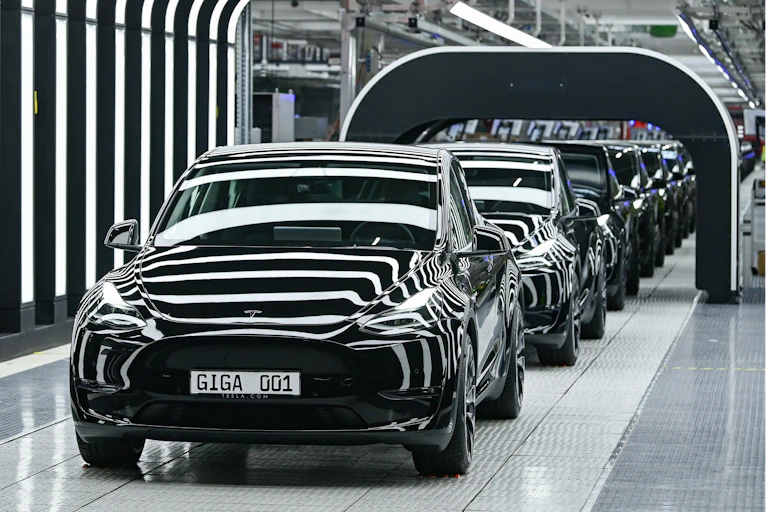 Several Tesla Model Y electric vehicles stand on a conveyor belt at the opening of a Tesla Gigafactory. Lighting creates a striped effect as the vehicles pass under a black arch.