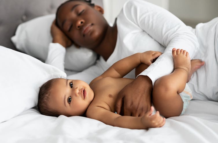 A baby lying on a bed is held by a woman lying next to him
