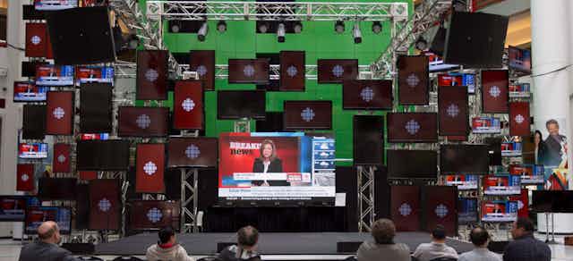People in a room watching CBC news, logos for the corporation are seen on several TV screens
