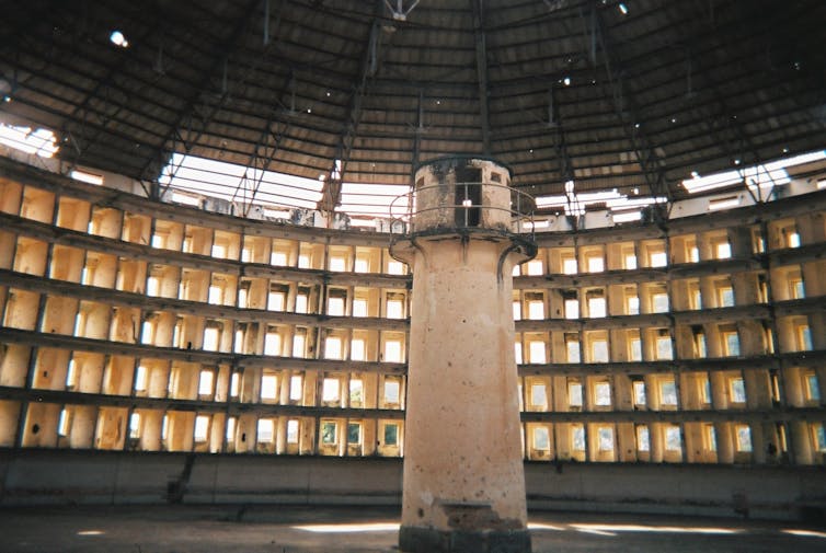 The interior of an empty panopticon prison, a circular, concrete room with a guard tower in the centre and dozens of cells with sunlight shining through