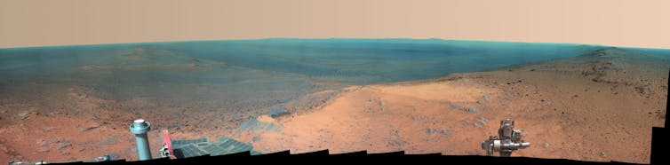 A false-color photo from the point of view of a rover standing at the cliff overlooking a brown, sandy desert-like area that looks blue in the distance.