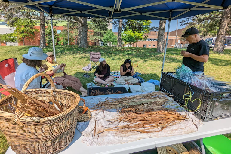 A group of people seen standing and sitting by a table weaving.