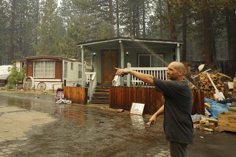 A main points off camera as sprinklers run, leaving wet ground in front of a row of mobile homes. The neighborhood is up against thick pine forest.