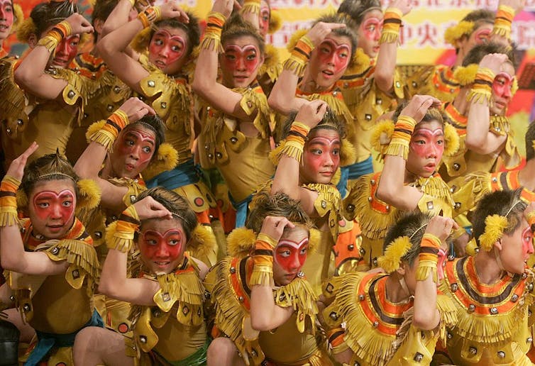 A dozen children in bright gold costumes and red face paint pose in a dance formation.