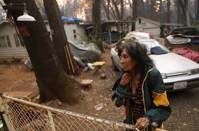 An older woman stands next to a fence of a home that survived the fire.