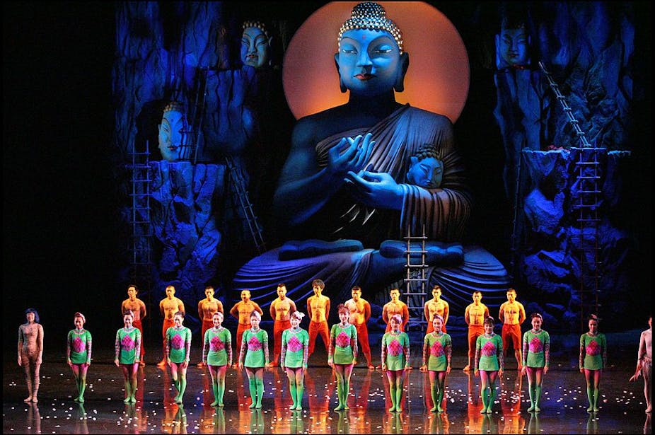 Two rows of performers in neon costumes on stage in front of a huge, blue statue of the Buddha.