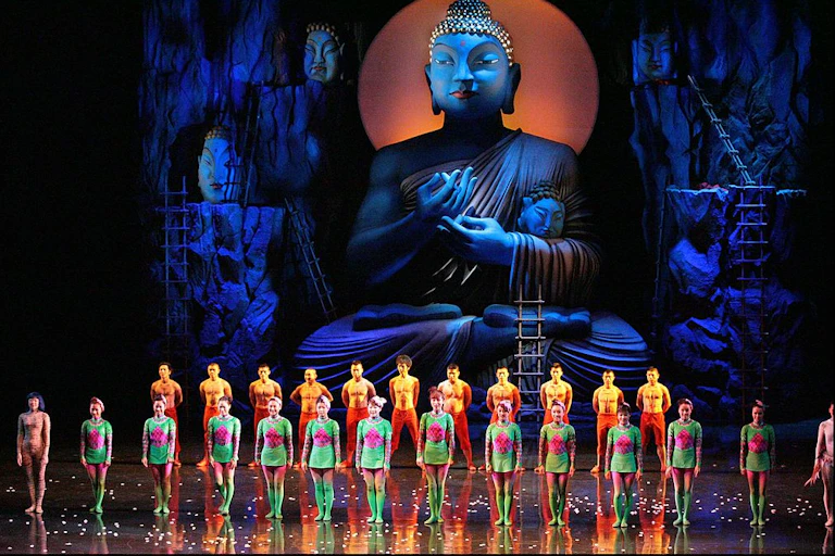 Two rows of performers in neon costumes on stage in front of a huge, blue statue of the Buddha.
