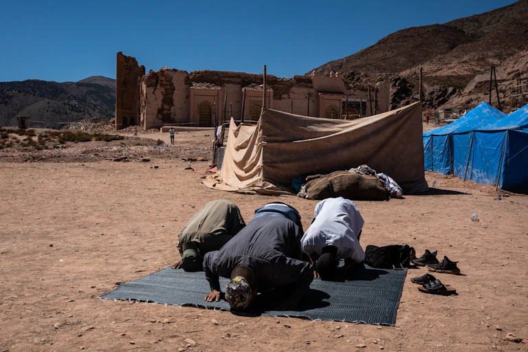 Three people prostrating in prayer on a mat. In the backdrop are mountains and a damaged ancient brick structure.
