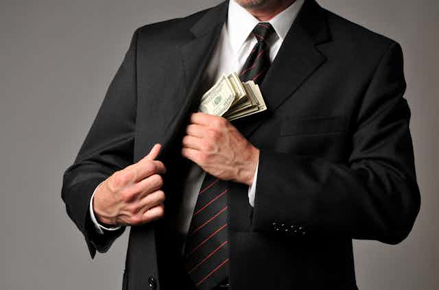 Standing before a gray background, a businessman slips a stack of hundred-dollar bills into his suit coat pocket. His face is out of frame.