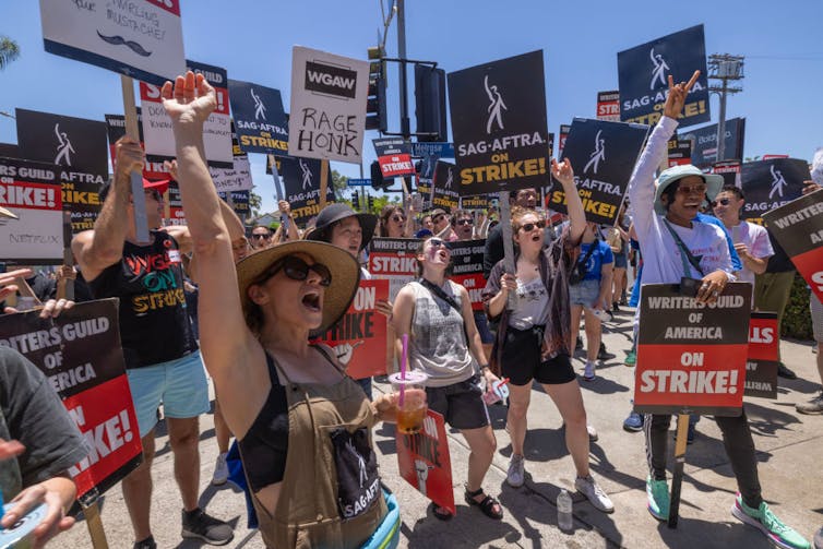 A group of striking workers yell, hold signs and thrust their arms skyward.