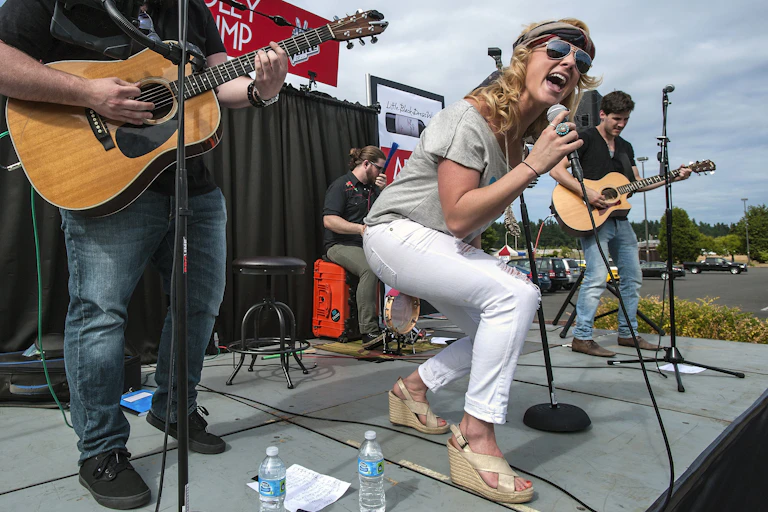 Woman wearing sunglasses flanked by two guitarists sings into microphone on stage.