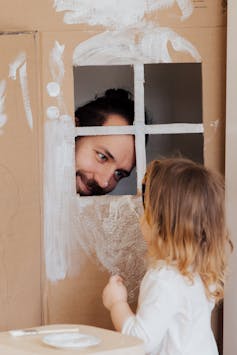 A man peeks in the window of a cardboard cubby with a young child inside.