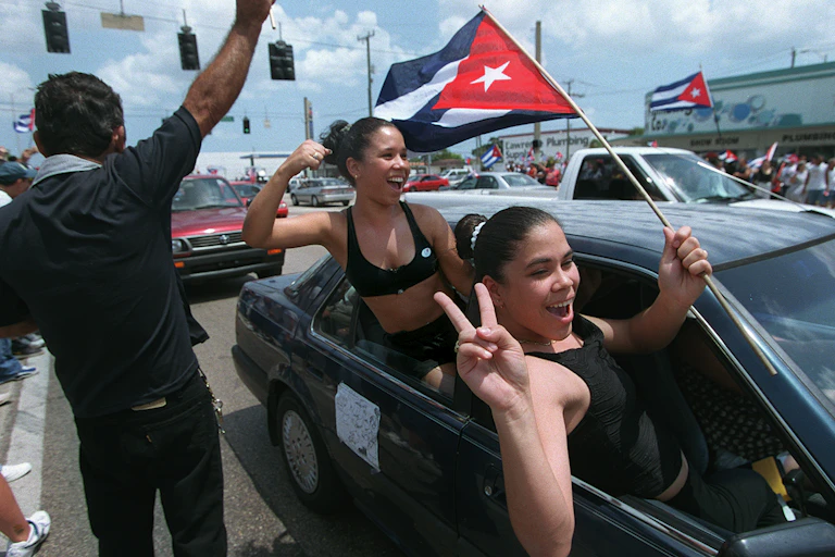 Dos jóvenes sonrientes se asoman a las ventanillas de los coches mientras una ondea una bandera cubana.
