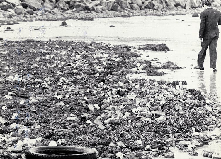 Black and white photograph of a man walking along a beach covered in discarded drink cans and other rubbish