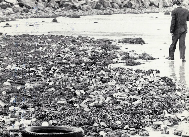 Black and white photograph of a man walking along a beach covered in discarded drink cans and other rubbish