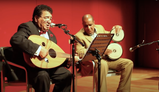 Two men sit on a stage in front of a red curtain. One plays a guitar-like instrument and sings into a microphone, another plays traditional Somali drums.
