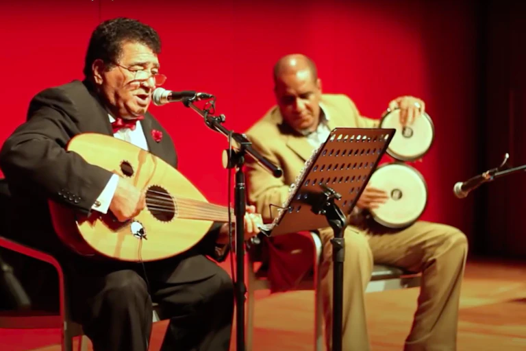 Two men sit on a stage in front of a red curtain. One plays a guitar-like instrument and sings into a microphone, another plays traditional Somali drums.