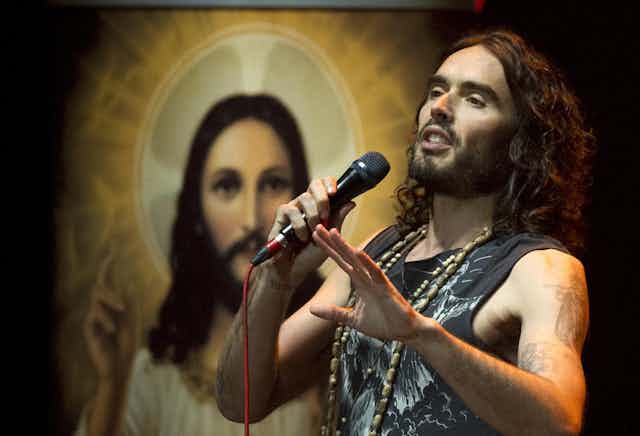 Russell Brand on stage holding a microphone in front of an image of Jesus.