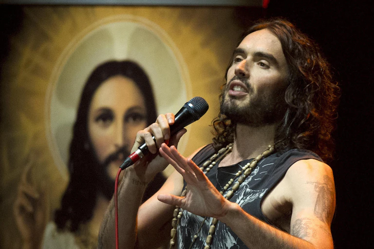 Russell Brand on stage holding a microphone in front of an image of Jesus.