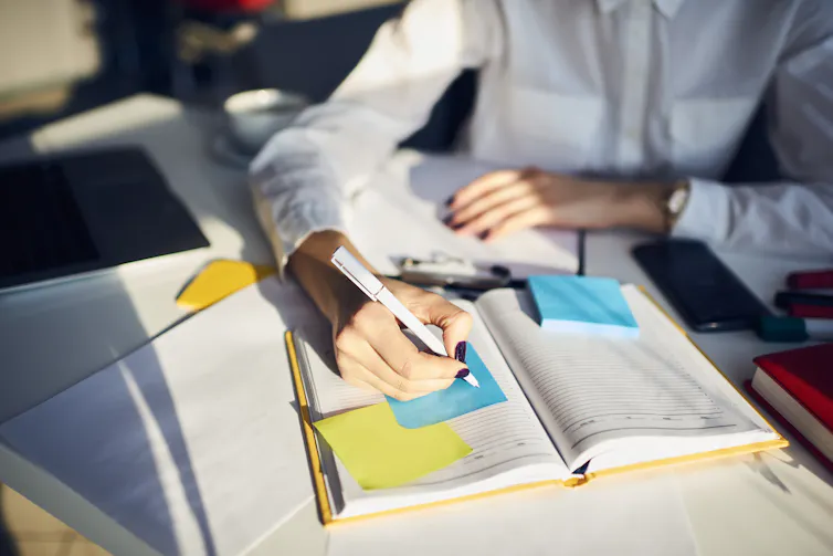 Photo of a woman's arms, writing in a notebook and on post-it notes on a desk
