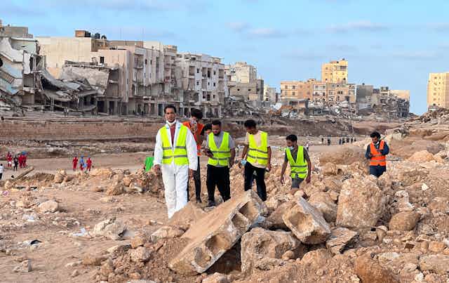 Men in high-visibility vests search rubble with crumbling buildings behind them.