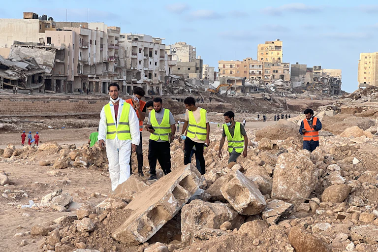 Men in high-visibility vests search rubble with crumbling buildings behind them.