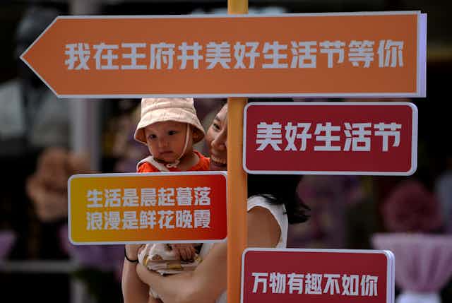 Mulher chinesa segura bebê no colo, por trás de placas de orientação escritas em chinês
