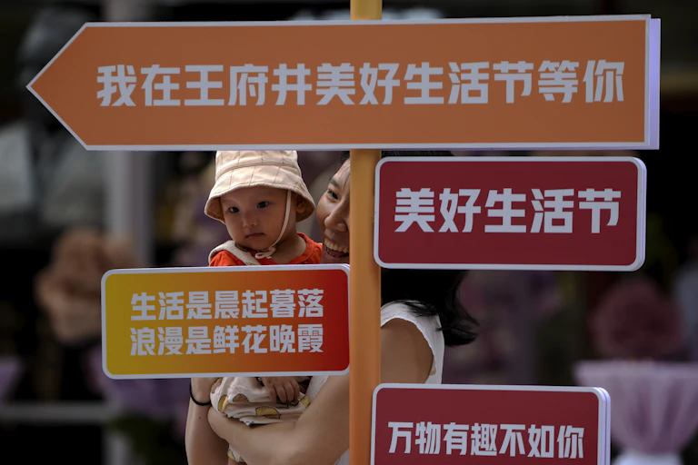 Mulher chinesa segura bebê no colo, por trás de placas de orientação escritas em chinês