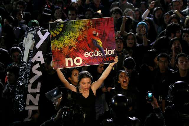 A woman in a tank top sorrounded by protesters holds up a colourful sign.