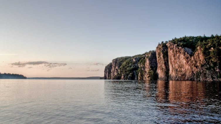 A high rock face topped with trees seen next to reflective water.