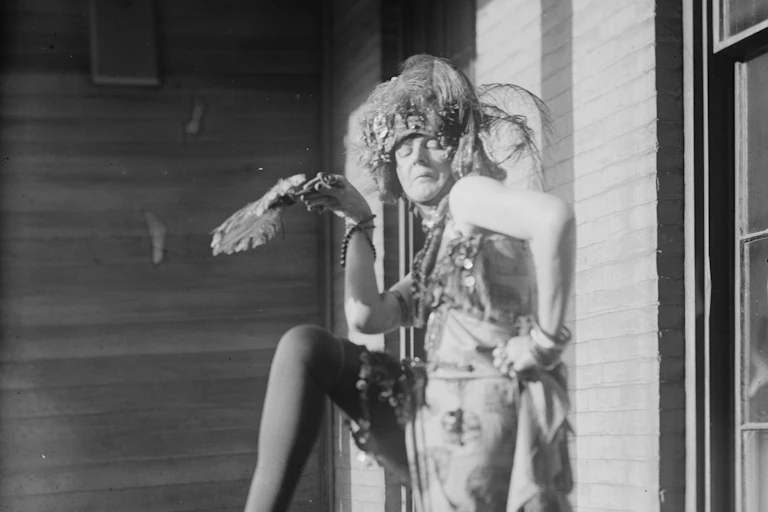A woman seen standing outdoors in a dramatic pose holding what appears to be a feather fan wearing a 1920s dress.