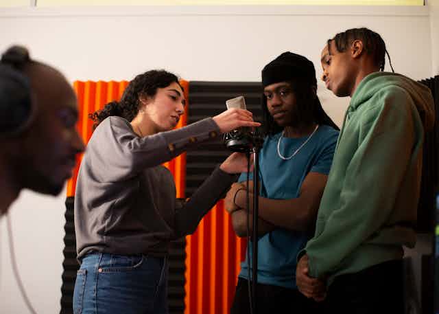 Woman adjusting microphone while rappers stand in studio. 