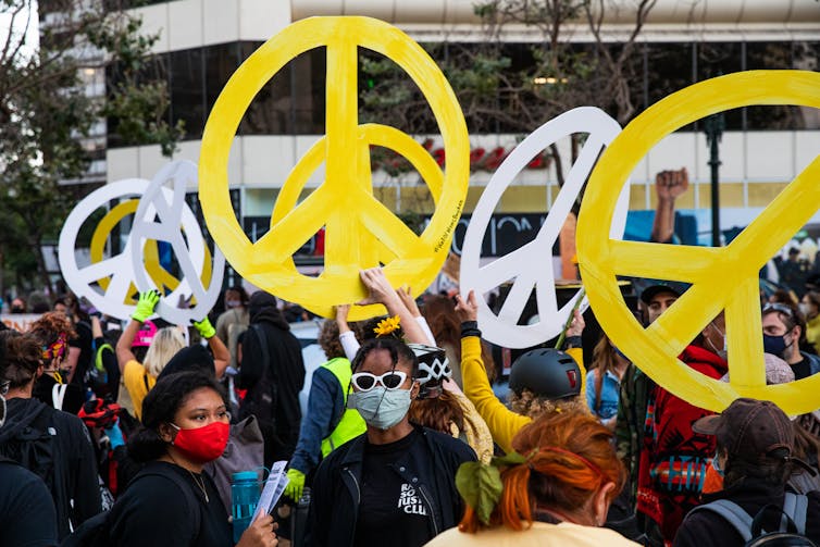 People wear face masks and hold large yellow and white peace signs on a city street.