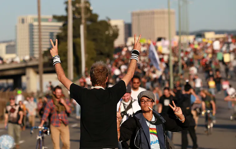 The back of a man flashing two peace signs with his hands is seen on a city street, with many other people walking past him.