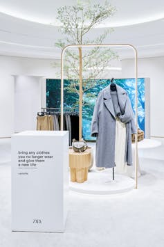 White store background with sales display of grey coat, tree and light behind white clothing collection bin.
