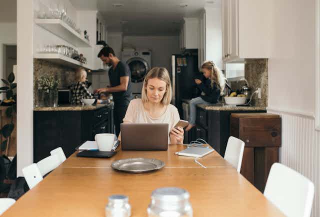 woman sits at dinner table in front of laptop with phone in hand while kids sit on counter and man stands near them in the kitchen behind her
