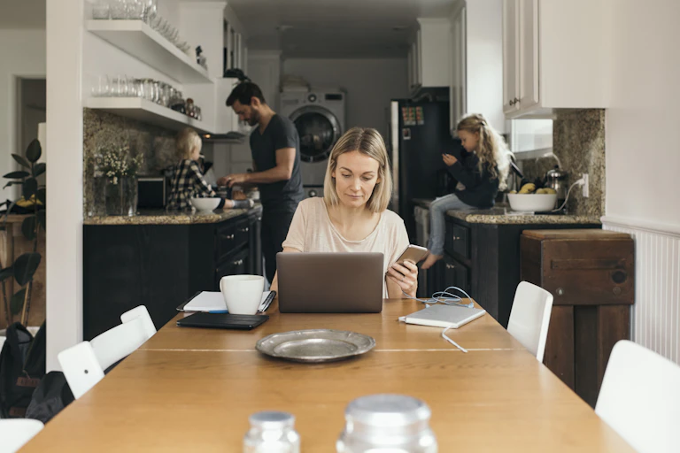 woman sits at dinner table in front of laptop with phone in hand while kids sit on counter and man stands near them in the kitchen behind her
