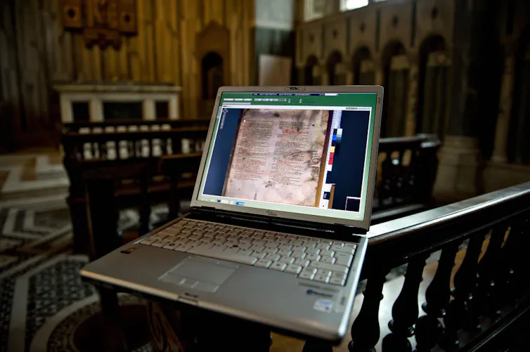 The screen of an open laptop, positioned in a dark church, shows a page of a very old-looking manuscript.
