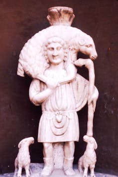 A light-colored carving of a man in a tunic holding a large sheep, with two smaller sheep at his feet.
