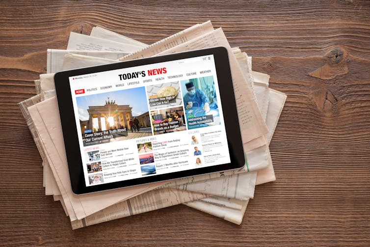 A tablet displaying a news page placed on top of a stack of newspapers